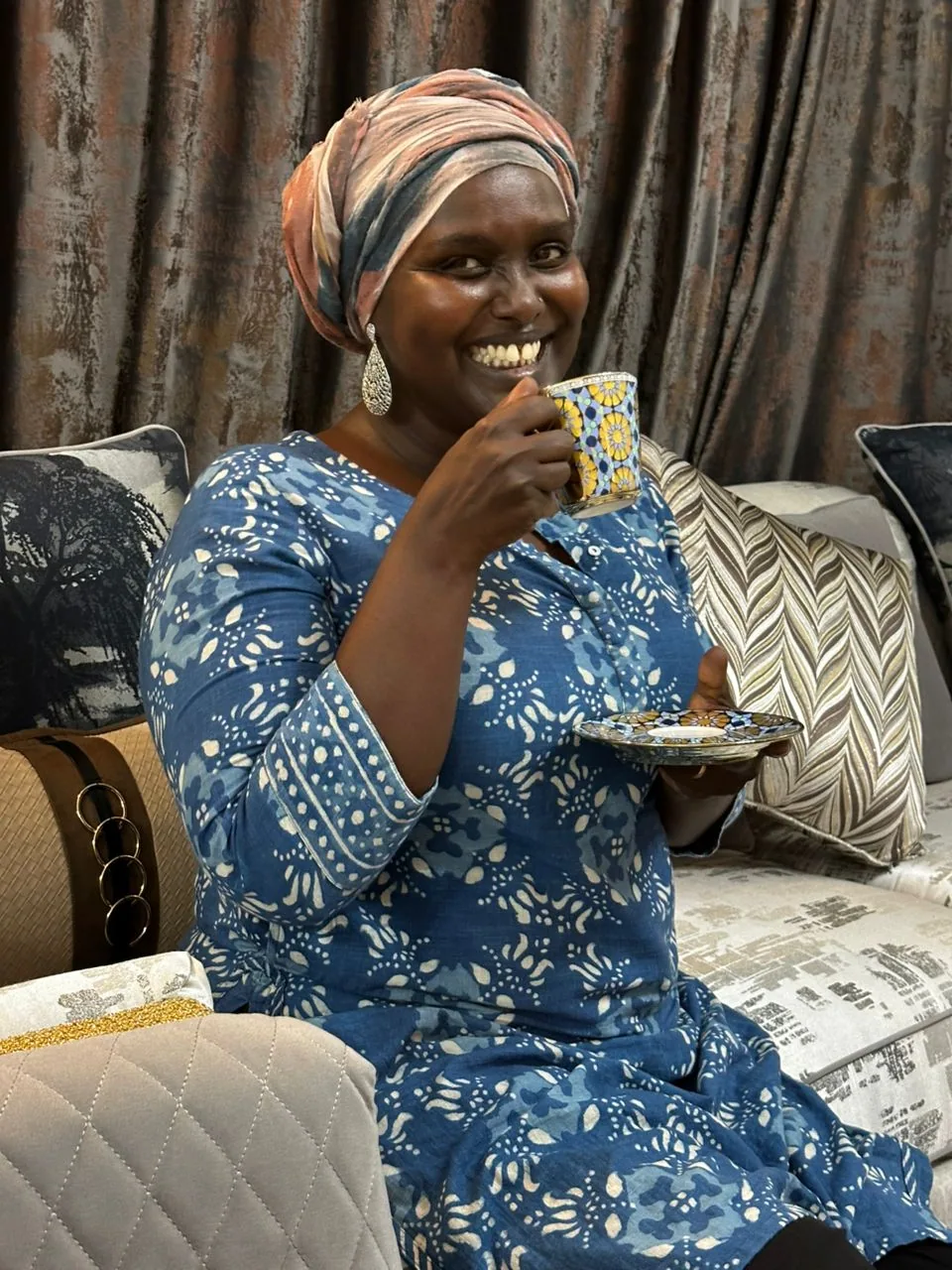 Gassira Tanaba souriante, une tasse de café à la main, dans un salon aux tissus imprimés